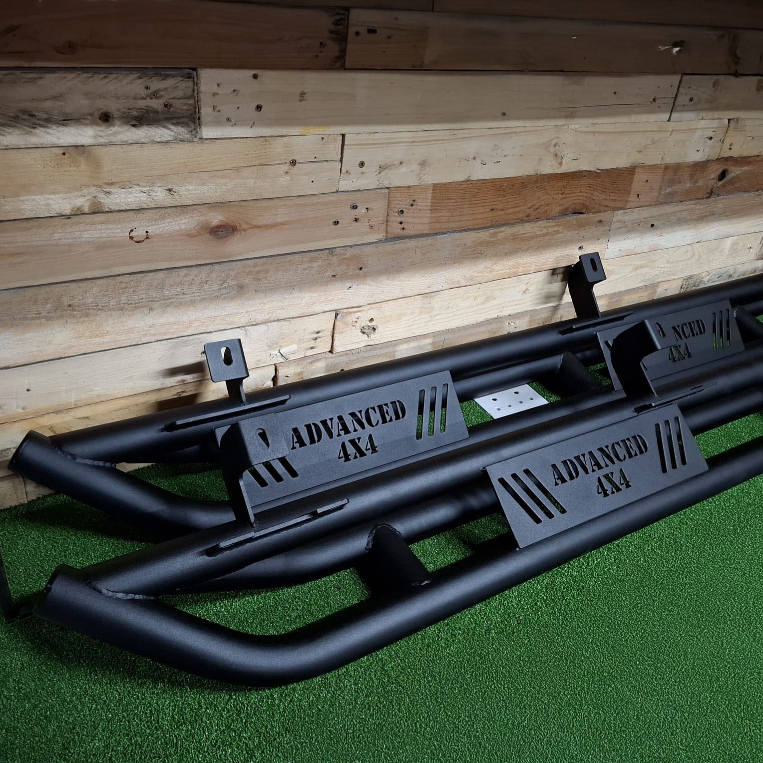 A pair of black steel rock sliders for a Jeep JK 5 Door vehicle, displayed against a wooden backdrop.